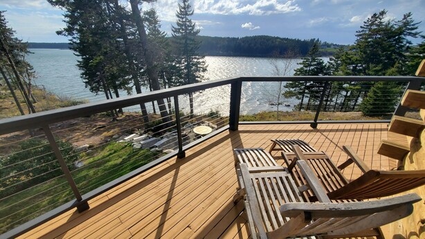 A wooden deck with two wooden chairs, a metal railing, and a view of a lake surrounded by trees. - Sunset Cabin by the Sea: Log cabin w/ stunning ocean views. Unwind in paradise! (Roque Bluffs)