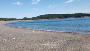 A pebbly beach with calm water, a forested hill in the background, and a clear sky. - Sunset Cabin by the Sea: Log cabin w/ stunning ocean views. Unwind in paradise! (Roque Bluffs)