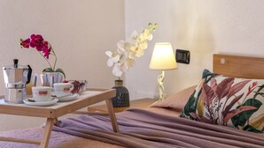 Una mesa de desayuno ordenada con cafetera, tazas y flores. También se ve una cama con un cojín floral y una lámpara.