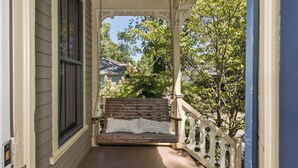Un portico con una panchina in legno, una ringhiera bianca con decorazioni e vista su una zona residenziale con alberi e case.