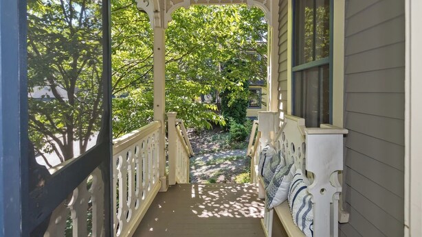 Un portico riparato con ringhiera in legno, una panca con cuscini e vista su un giardino con alberi e una casa sullo sfondo.