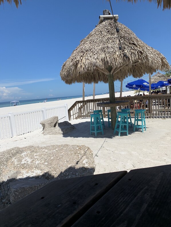 Une terrasse au bord de la plage, dotée d’un parasol en chaume, d’une plateforme en bois et de tabourets turquoise.