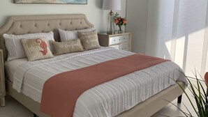 A bedroom with a large bed, a nightstand with a lamp, a painting of a beach, and a window with curtains. - Our house at the beach Most see<br>At siesta key  (Siesta Key)