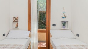 A bedroom with two beds, a wooden door, a view of greenery outside, and a small shelf with a plant. - Vacation home Cisto by Interhome (Costa Paradiso)