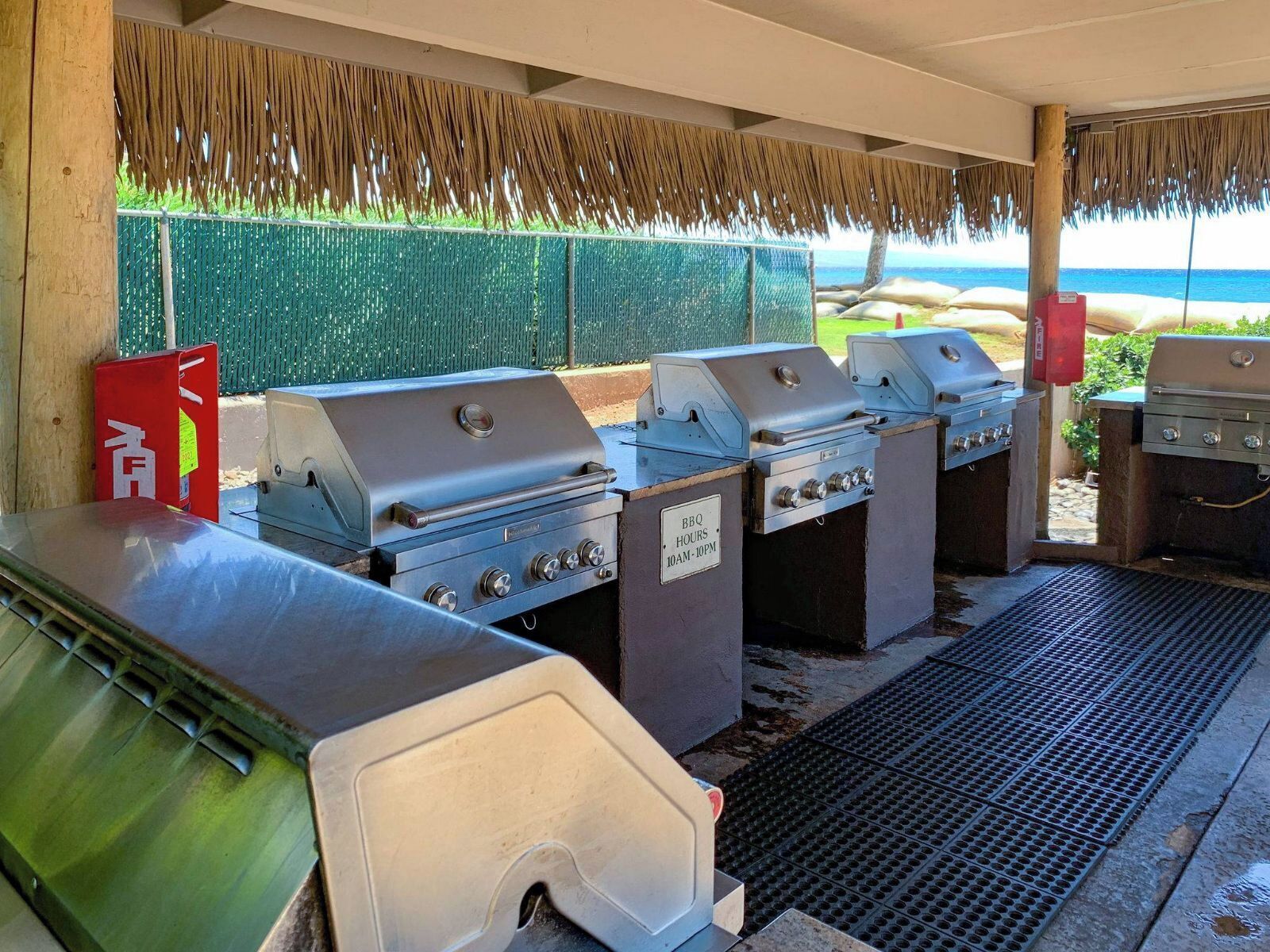 An outdoor barbecue area with multiple grills, a red fire extinguisher, and a sign indicating BBQ hours.
