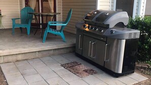 Een terras buiten met een barbecue, turquoise stoelen en een tafel.