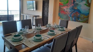 Een eetruimte met een lange tafel gedekt voor zes personen, een flatscreen televisie aan de muur en een kleurrijk abstract schilderij.