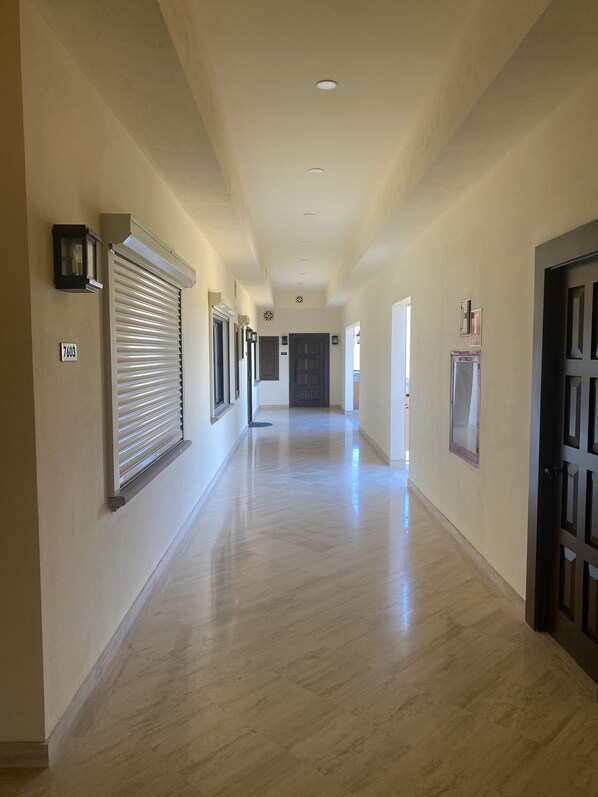 Un pasillo largo y estrecho con piso de color claro, una puerta a la derecha y una ventana con persianas a la izquierda.