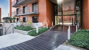 Un bâtiment moderne avec une façade en briques, des balcons et un chemin en bois menant à une entrée en verre.