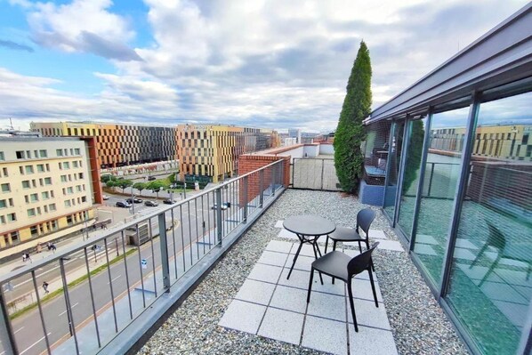 A rooftop terrace with a view of the city, featuring a glass balustrade, a seating area with two chairs, and a tall tree.