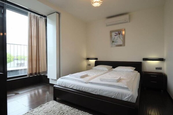 A bedroom with a large bed, two bedside tables, a window with curtains, and a wall-mounted air conditioner.