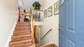 Una scala in legno conduce a un secondo piano con una porta blu, un tappetino d'ingresso e stampe botaniche incorniciate alle pareti.