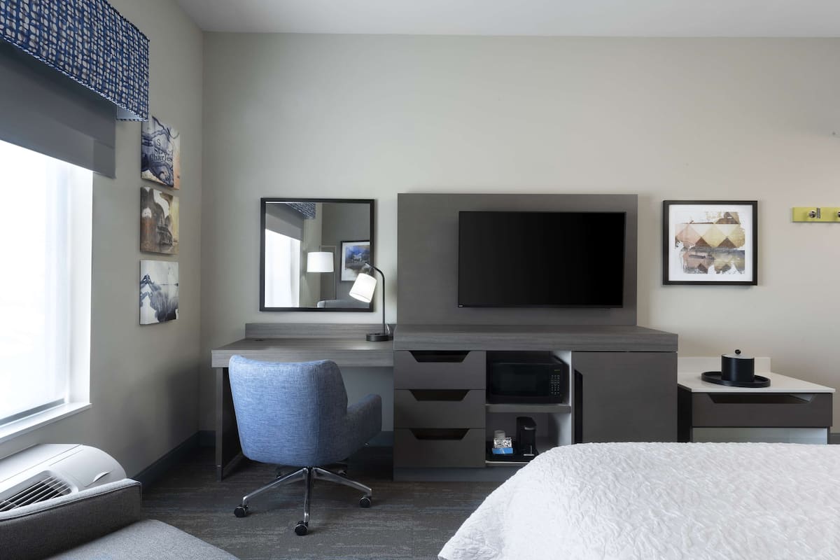A hotel room with a bed, a desk with a chair, a flat-screen TV, a microwave, and a small bedside table.