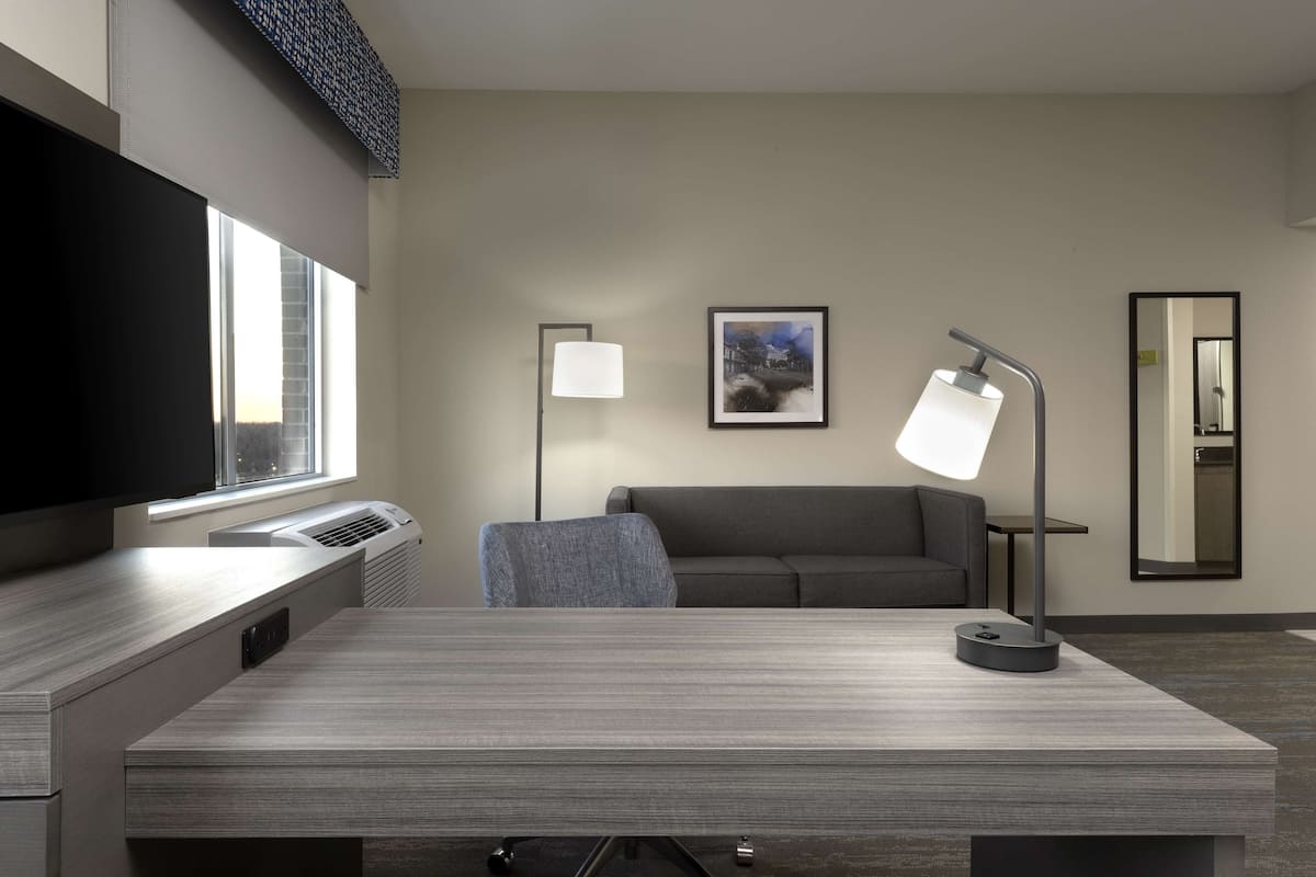 A modern hotel room with a flat-screen TV, a desk with a lamp, a sofa, and a large mirror.