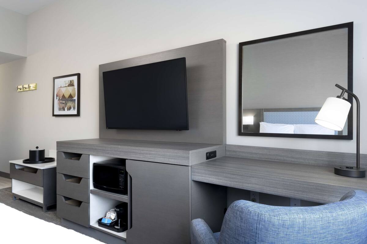 A modern hotel room with a flat-screen TV mounted on the wall, a small bedside table with a lamp, a chair, and a built-in microwave.