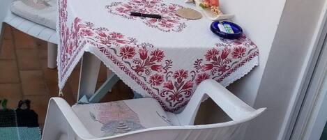 A small outdoor seating area with a table covered by a floral tablecloth, white plastic chairs, and a green and white chair leaning against the wall.