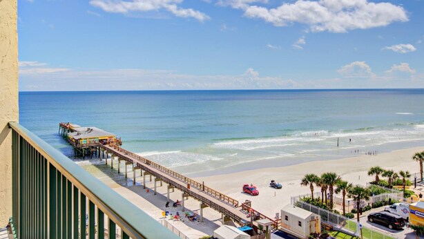 A beach view with a pier extending into the ocean, a sandy beach, and a few parked cars.