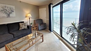 A living room with a sofa, a glass coffee table, a patterned armchair, a lamp, and a view of the sea through large windows.