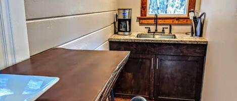 Cocina compacta con una mesa de madera, armarios oscuros, una ventana con vistas a árboles y una cafetera en la encimera.