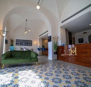 Um lobby com sofá verde, azulejos estampados no chão, balcão de recepção e uma pintura emoldurada com vista para a cidade.