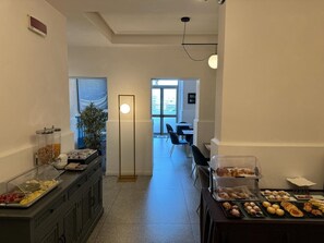 Eine Hotelhalle mit einem Buffetbereich, der verschiedene Speisen anbietet, sowie einer Sitzecke mit Tischen und Stühlen, und einem großen Fenster, das für natürliches Licht sorgt.