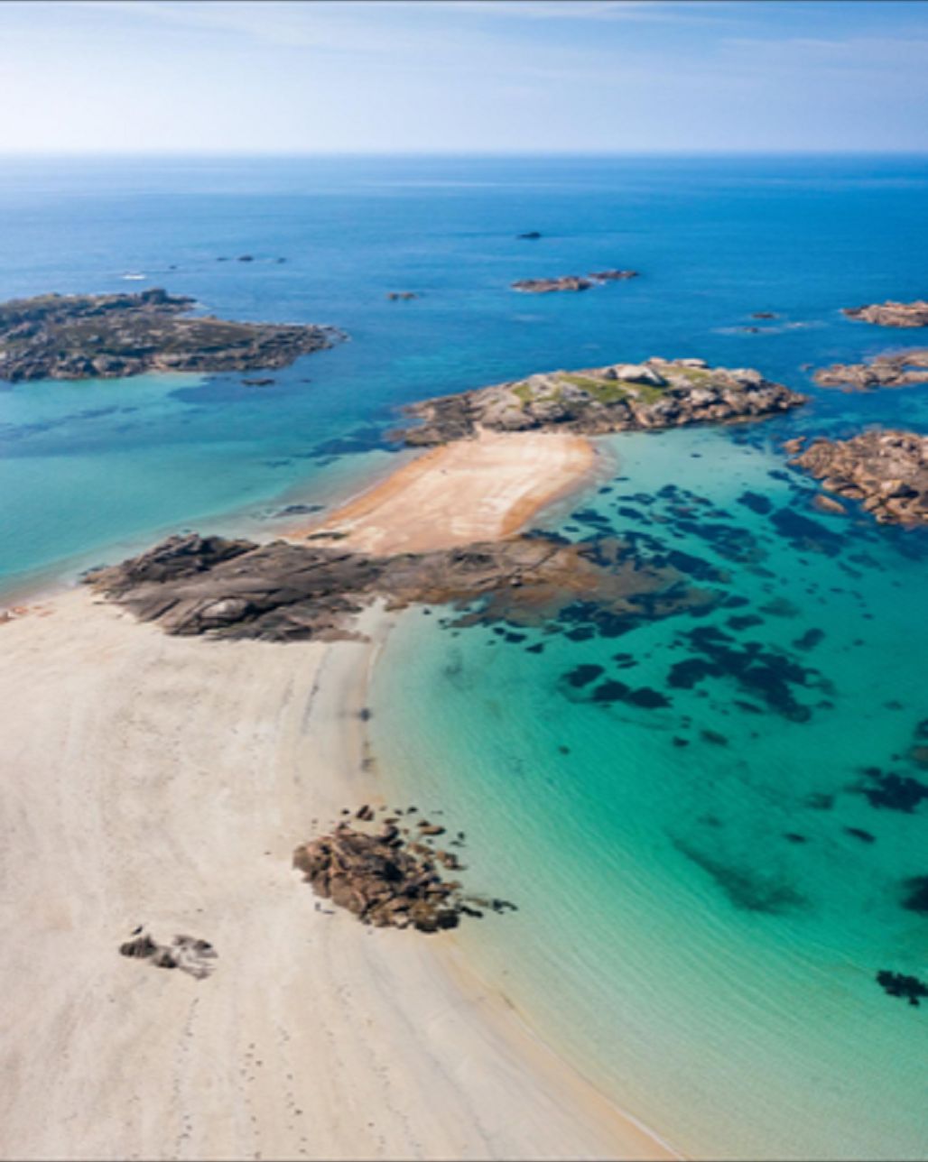 An aerial view of a coastal area with clear turquoise waters, sandy beaches, and rocky outcrops.