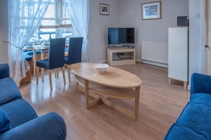 A living room with a wooden dining table, blue chairs, a TV on a stand, and a white cabinet.