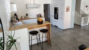 A modern kitchen with a central island, two bar stools, a fruit bowl, and a refrigerator.