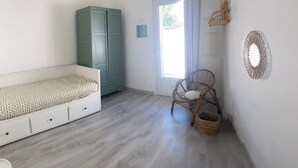 A room with a bed, a wardrobe, a wicker chair, a small basket, and a wall-mounted device.