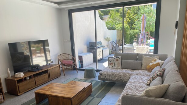 A modern living room with a flat-screen TV, wooden furniture, a grey sofa, and a view of the outdoors.