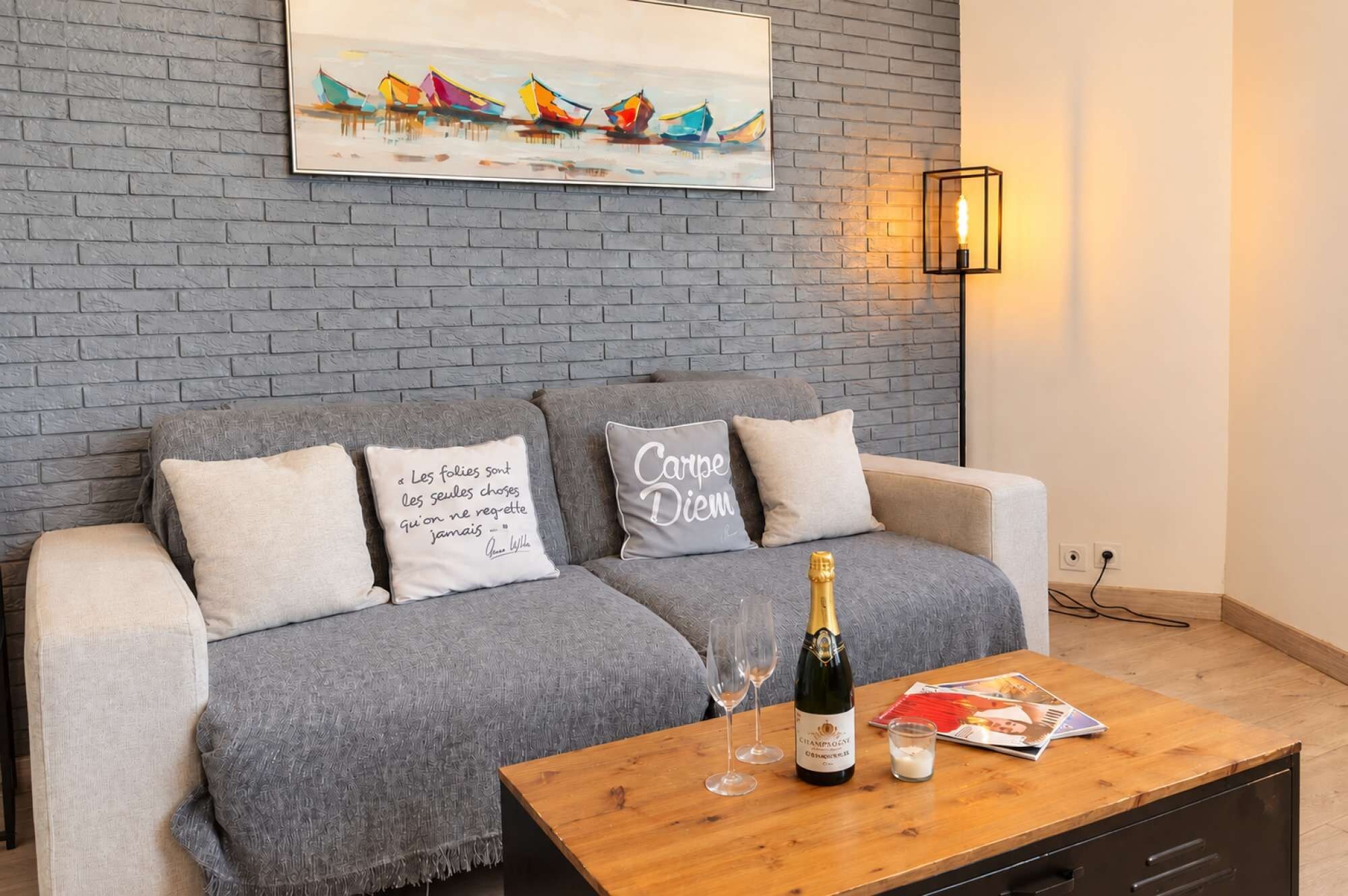 A modern living room with a grey brick wall, a grey sofa with decorative pillows, a wooden coffee table with a bottle of champagne, glasses, and magazines.