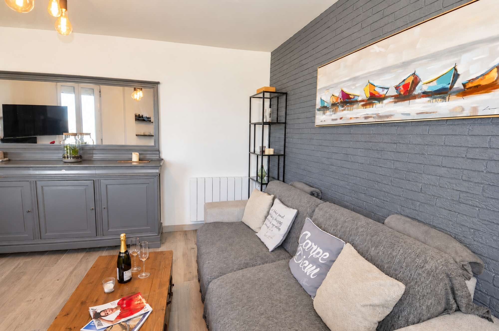 Ein Wohnbereich mit grauem Sofa, einem Couchtisch mit Flasche und Gläsern, einem Gemälde über dem Sofa und einer Küche im Hintergrund.
