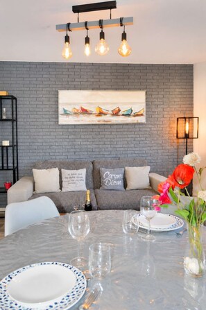 A dining area with a brick wall, a painting of boats, a sofa with cushions, and a table set for two with wine glasses and plates.