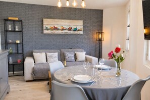 A modern living room with a grey brick wall, a sofa with decorative pillows, a dining table set for two, and a painting above the sofa. - Apartment near port center and beach - Sea view (Courseulles-sur-Mer)