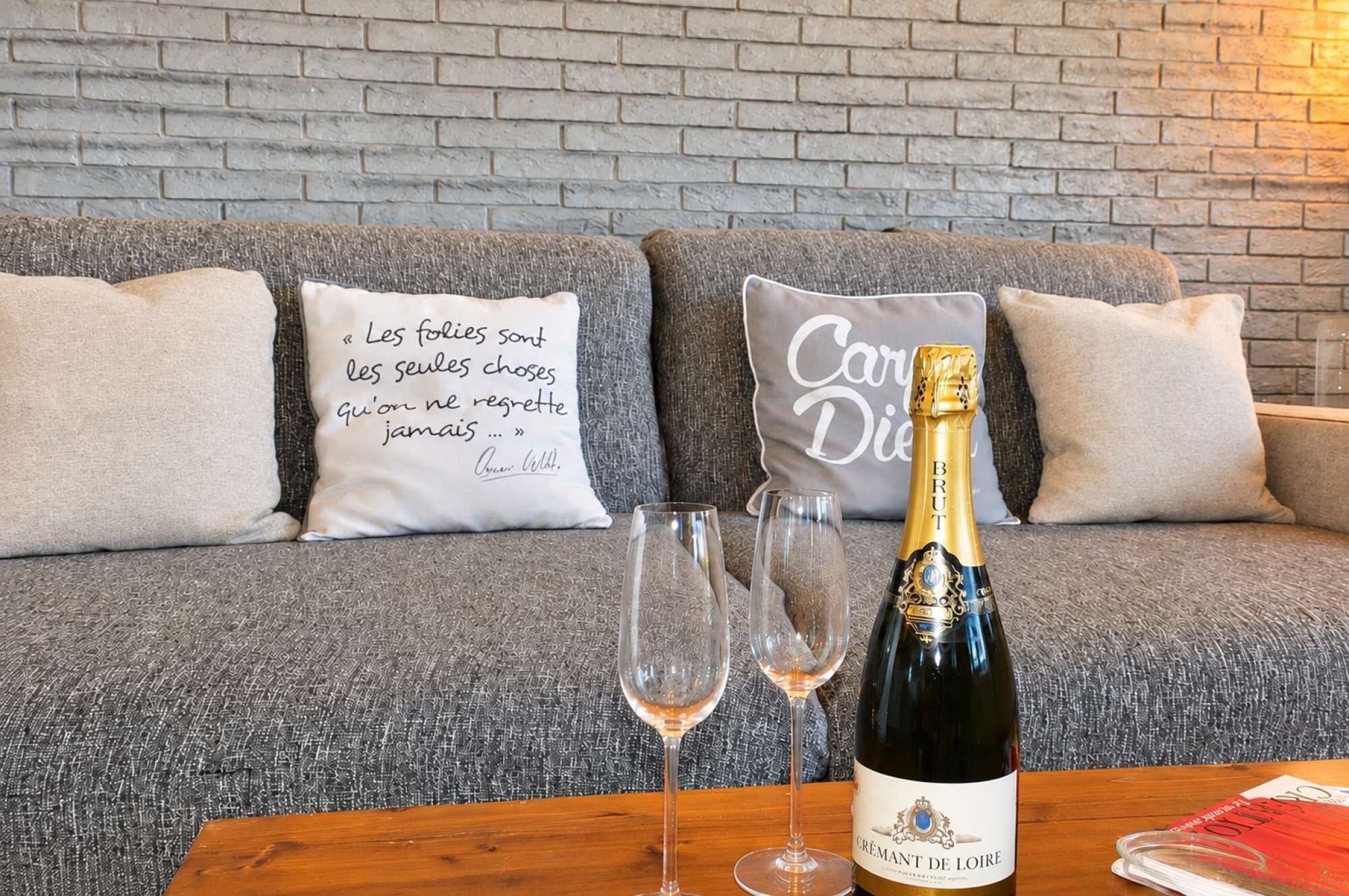 En grå sofa med to puder, hvoraf den ene har et citat og den anden et navn, en flaske champagne og to glas på et træbord.
