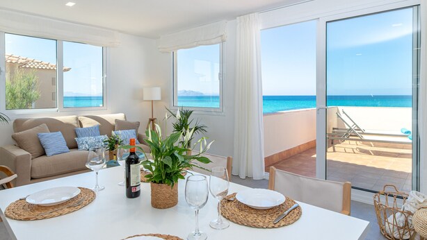 Een zithoek met een witte tafel voor vier personen, een bank met kussens, een kamerplant en uitzicht op het strand door grote ramen.