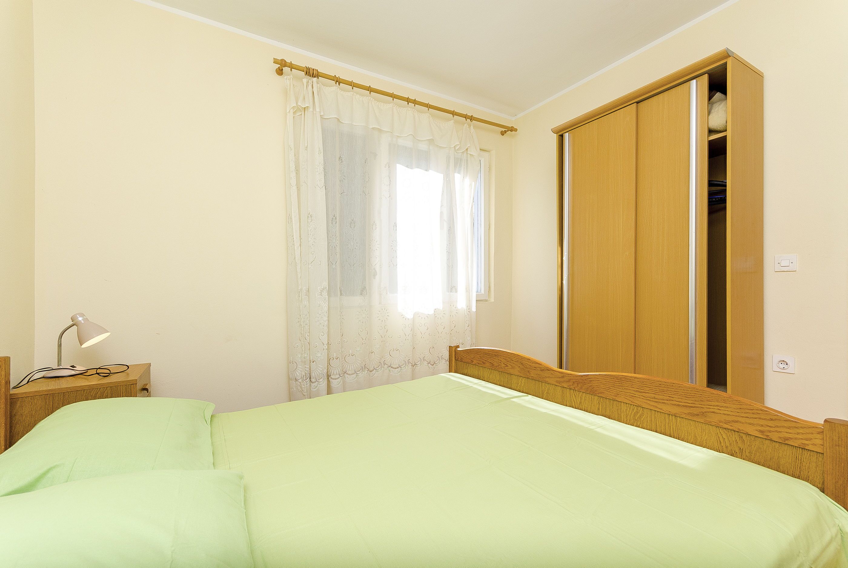 A bedroom with a bed, a nightstand with a lamp, a window with curtains, and a wardrobe.