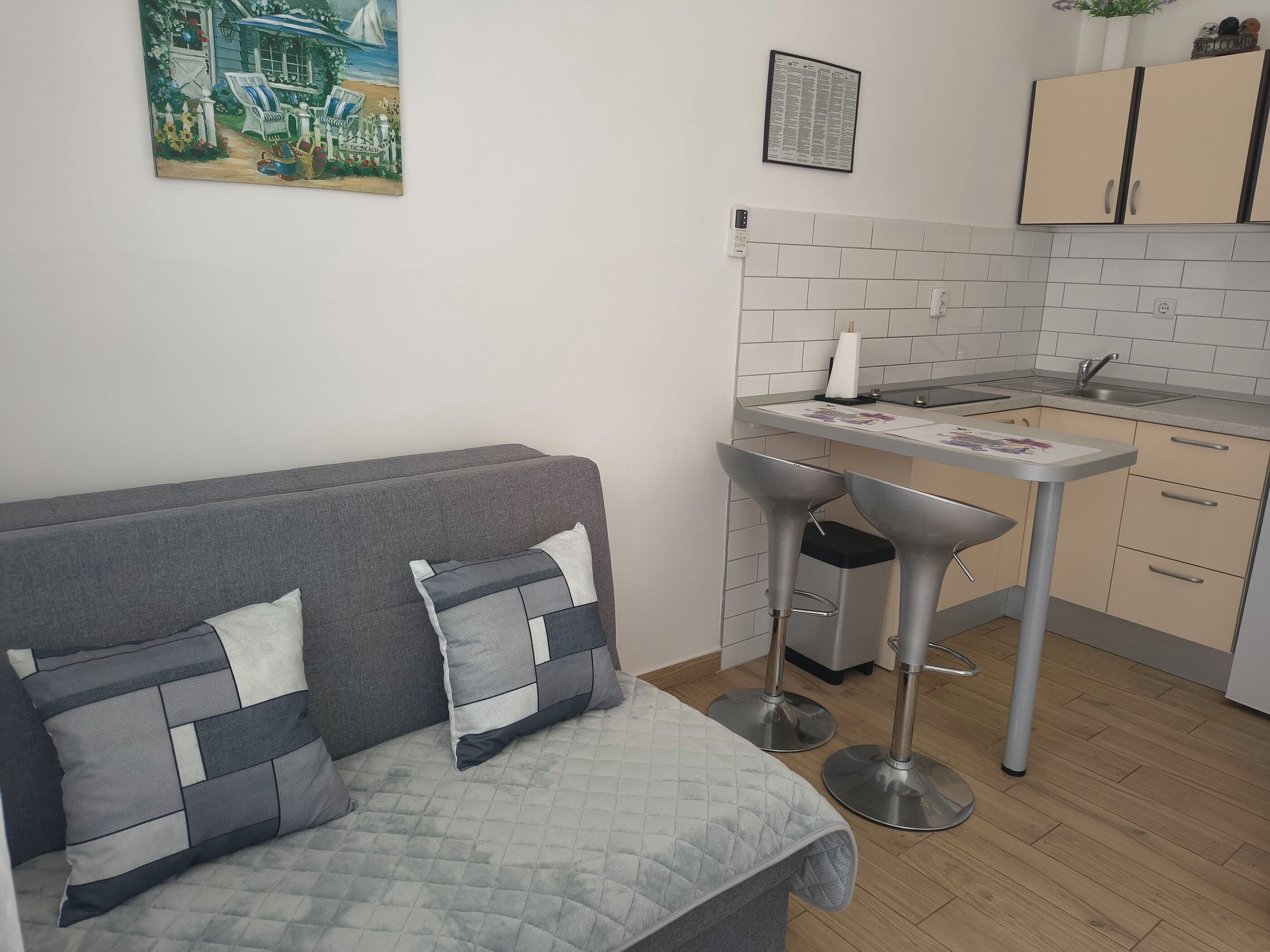 A modern kitchen with a grey sofa, two bar stools, a small table, and a tiled backsplash.