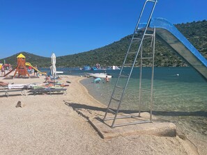 A beach with a playground, clear water, and boats. - Apartments Matas (Rogoznica)
