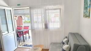 A room with a grey sofa, a wooden table with a chair, a window with curtains, and a door leading outside. - Apartments Matas (Rogoznica)