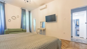 Ein Schlafzimmer mit einem Bett, einem grünen Sofa, einem Fernseher und einem durchgehenden Türrahmen einsehbaren Badezimmer.