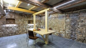 Ein rustikaler Raum mit Steinwänden, einem Holztisch und -stühlen, einem Steinschränkchen mit Flaschen und einer schrägen Decke mit freiliegenden Balken.