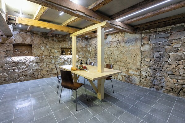 A rustic stone-walled room with a wooden table and chairs, a stone shelf with bottles, and a sloped ceiling with exposed beams. - House Goldie (Tribalj)