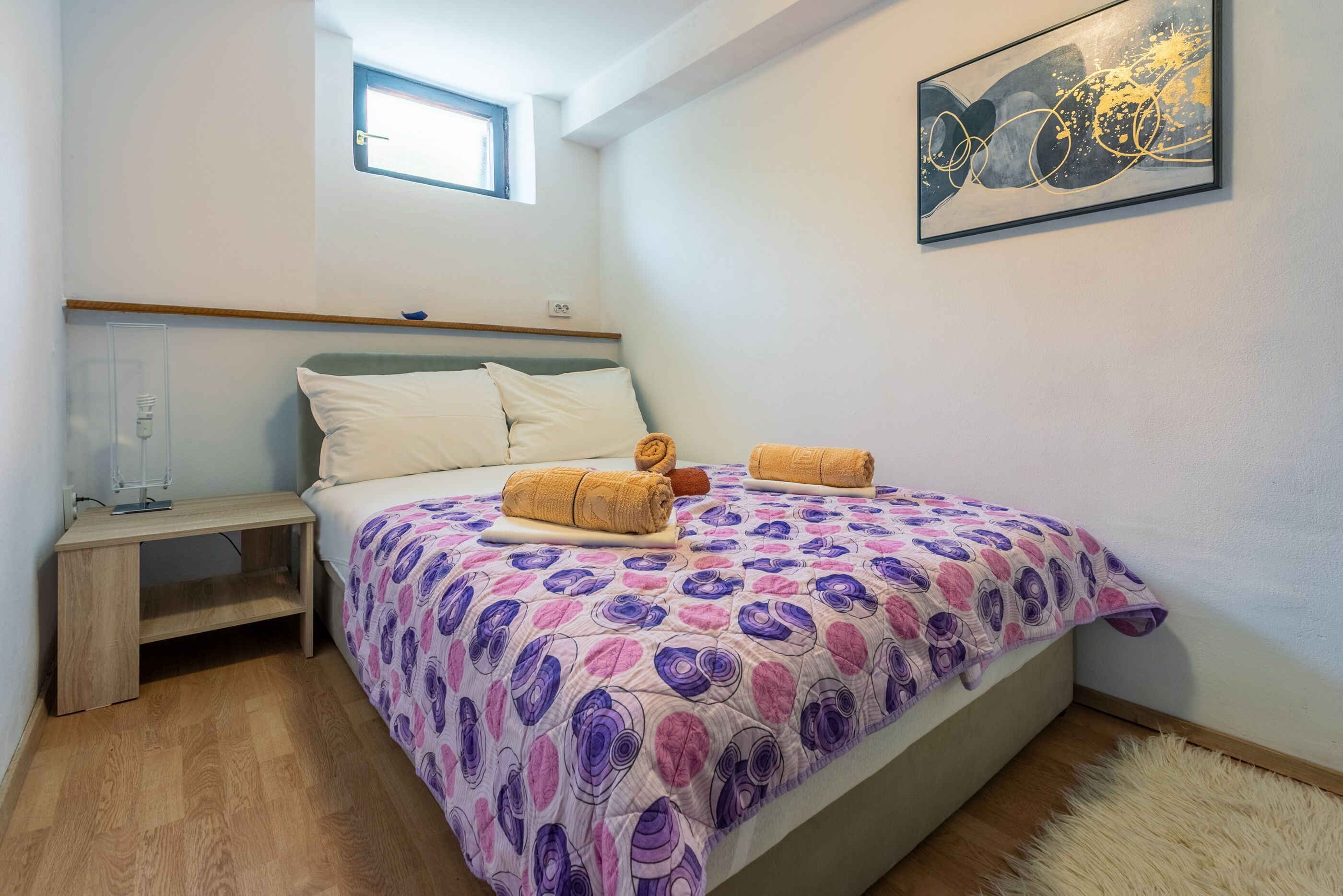 A bedroom with a bed, a nightstand, a window, and a painting on the wall.