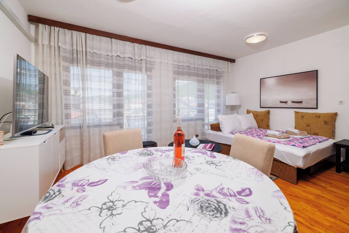 A modern hotel room with a round dining table, a bed with a floral bedspread, a desk with a computer, and a large window with sheer curtains.