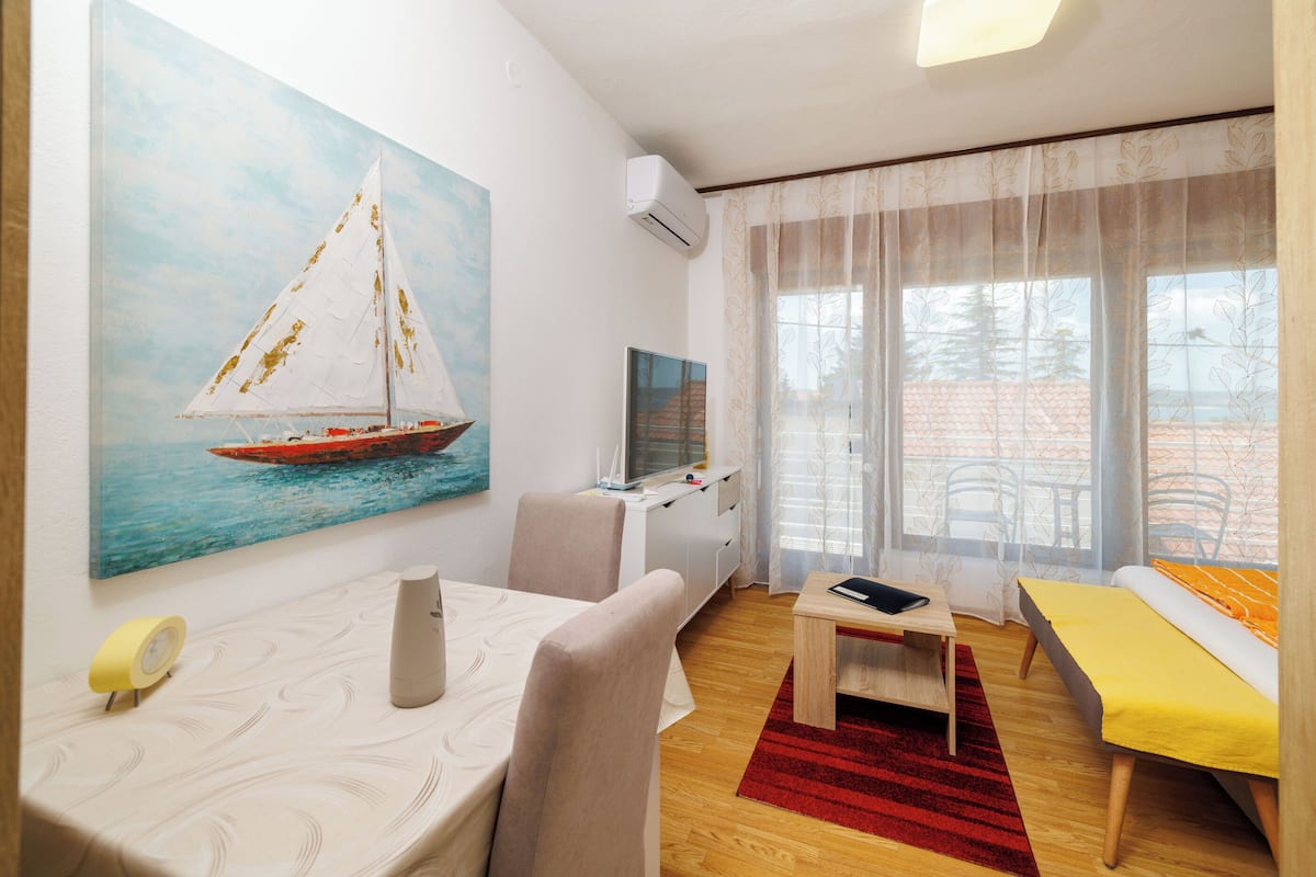 A modern living room with a sailboat painting, a dining area with a table and chairs, a sofa, and a large window with a view.