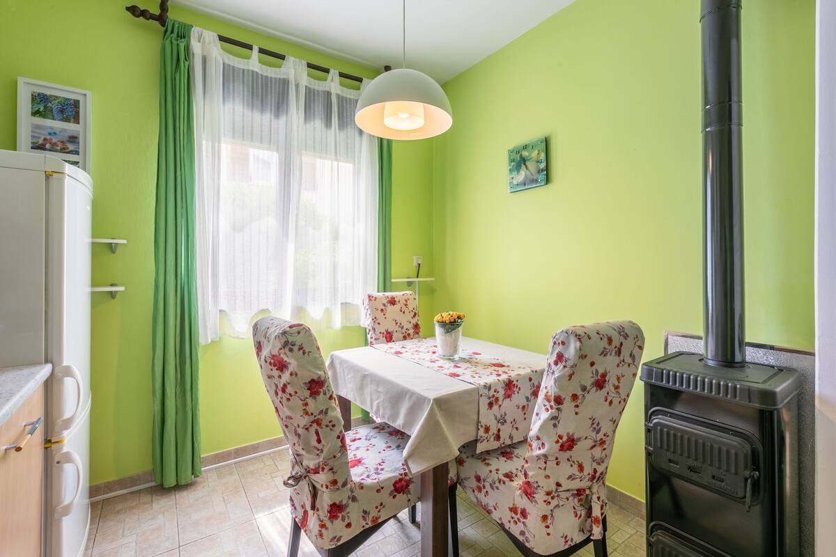 A dining area with a table set for two, chairs with floral upholstery, a white tablecloth, a green wall, a white ceiling light, a black stove, and a window with sheer curtains.