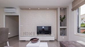 A modern living room with a flat-screen TV on a white console, a large grey sofa, and a white coffee table. - Apartments Dora (Okrug Gornji)