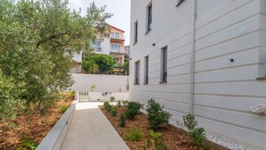 Ein schmaler Pfad, der von einem weißen Gebäude mit zahlreichen Fenstern und einem kleinen Garten mit verschiedenen Pflanzen und Sträuchern gesäumt wird.
