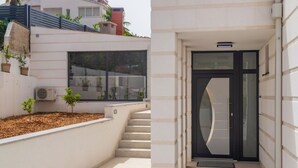 A modern building entrance with a glass door, a small outdoor area with plants, and a staircase leading to the entrance. - Apartments Dora (Okrug Gornji)